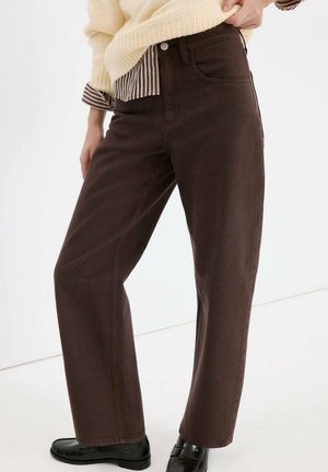 Femme portant un pantalon droit marron foncé associé à des mocassins noirs et un pull beige par-dessus une chemise rayée.