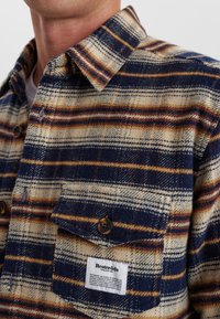 Camicia di flanella a scacchi in colori beige, blu navy e ruggine, con colletto button-down, taschino sul petto e etichetta in tessuto sulla tasca inferiore.