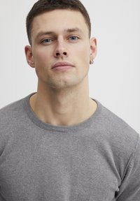 Grå strikkegenser med rund hals, tettsittende fasong og subtil tekstur. Modellen har kort brunt hår og en øredobb. Bakgrunnen er ensfarget.