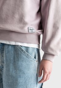 Felpa rosa chiaro con polsini e orlo a costine, dotata di etichetta. Indossata sopra una camicia bianca e abbinata a jeans denim blu chiaro.