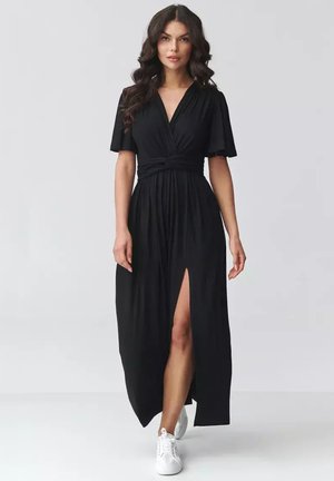 Kobieta w czarnej maxi sukience z dekoltem w serek, krótkimi rękawami, rozcięciem z przodu i białych trampkach, stojąca na tle jednolitego jasnego tła.