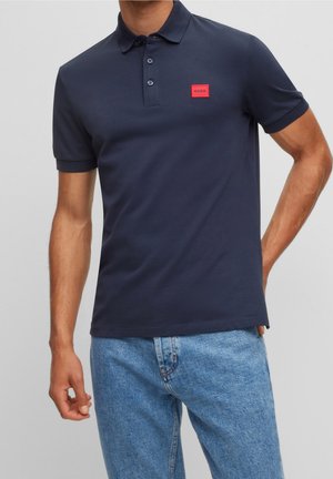 Polo azul marino de algodón con una botonadura de tres botones y un pequeño parche con logo rojo en el pecho, combinado con unos vaqueros azul claro.