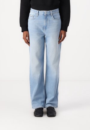 Personne portant un jean large bleu clair, un haut à manches longues noir, et des chaussures noires à bout carré, debout devant un fond blanc.