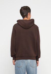 Brown hoodie with a large hood, relaxed fit, and ribbed cuffs. The fabric appears soft and textured, paired with light blue denim jeans.