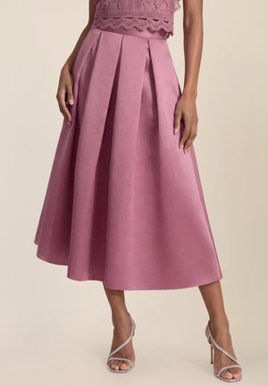 Frau trägt einen plissierten, altrosafarbenen Midi-Rock, ein Spitzentop und silberne, hochhackige Sandalen mit Riemchen und steht vor einem schlichten Hintergrund.