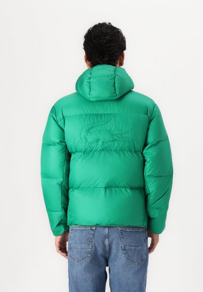 Doudoune verte avec capuche et logo gaufré dans le dos. Matière lisse, poignets côtelés et coupe décontractée. Portée avec un jean bleu.