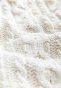 Mīksta balta adīta auduma ar izsmalcinātām kabeļu un ribas rakstu detaļām, radot teksturētu, mājīgu virsmu.