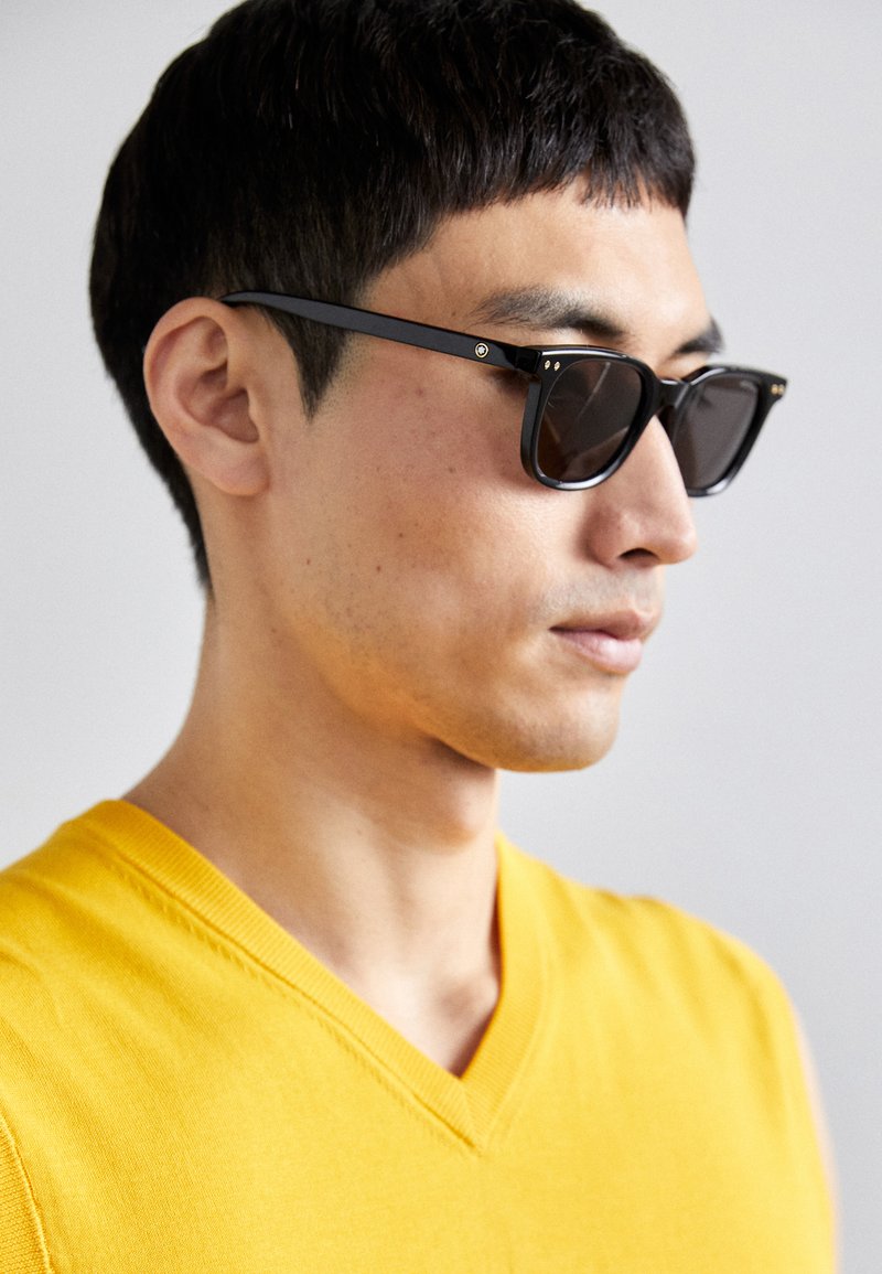 Aurinkolasit, joissa on mustat kehykset ja tummat linssit. Huomiota herättäviä yksityiskohtia ovat pienet kultaiset korostukset saranassa ja suorakulmainen muoto.