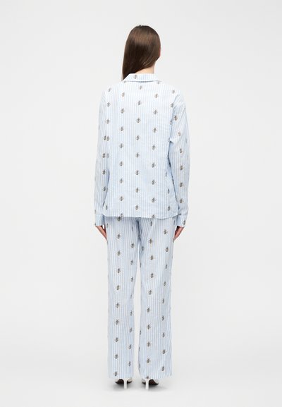 Woman standing facing away, wearing light blue and white striped pajamas with bee patterns and white heels against a plain white background.