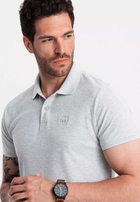 Camisa polo gris de manga corta, con un logo sutil en el pecho, combinada con un reloj negro con correa marrón. Textura de algodón suave.