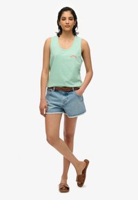 Kobieta stoi z rękami w kieszeniach, ubrana w zielony bezrękawnik, dżinsowe szorty z brązowym plecionym paskiem oraz brązowe sandały.