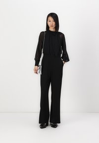 Zwarte jumpsuit met doorschijnende lange mouwen, hoge halslijn en een strikdetail. Gecombineerd met glanzende zwarte schoenen en een kleine metallic handtas.