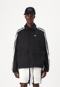 Casaco preto com riscas brancas, apresentando um design com colarinho, botões de pressão e dois bolsos frontais. Combinado com calções brancos e um boné.