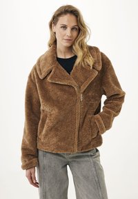 Vrouw met golvend blond haar draagt een bruine zachte ritsjas en grijze jeans, poseert met één hand in de zak tegen een witte achtergrond.