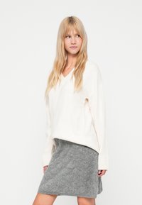 Svetlý krémový sveter s výstrihom do V a rebrovanými manžetami, v kombinácii s textúrovanou sivou sukňou. Oblečenie má uvoľnenú, vrstvenú siluetu.