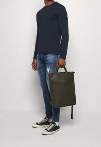 Bolso tote de piel verde con asas duales, forma rectangular, textura suave y bolsillo frontal. Combinado con jeans azules y zapatillas negras.