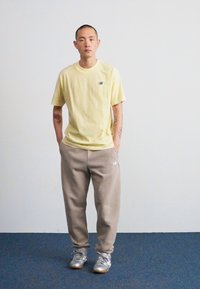 Geltona trumpa rankovė t-shirt su mažomis logotipu, derinama su šviesiai pilkais sportiniais kelnais ir pilkais sportiniais bateliais, stovint ant vientiso fono.