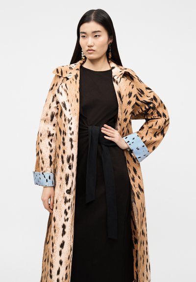 Νεαρή γυναίκα που φοράει ένα μακρύ, ταμπά με μαύρες βούλες παλτό με μπλε μανσέτες πάνω από ένα μαύρο φόρεμα δεμένο στη μέση, στέκεται με το ένα χέρι στη μέση της.