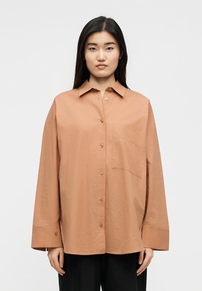 Γυναίκα με μακριά μαύρα μαλλιά, φορώντας μια χαλαρή, ταμπά πουκαμίσα με κουμπιά, τσέπη στο ύψος του στήθους και φαρδιά μανίκια, στέκεται μπροστά σε λευκό, απλό φόντο.