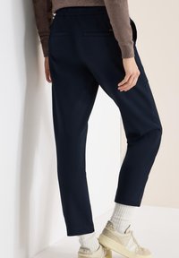 Pantalon bleu marine fuselé en tissu doux, avec taille élastique et deux poches arrière. Porté avec des baskets beige et des chaussettes crème texturées.