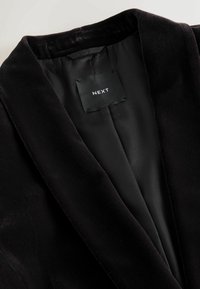 Čierny sametový blazer s hladkým saténovým podšívkou. Pozoruhodné rysy zahŕňajú klopový golier a štítok vo vnútri s logom značky "NEXT."