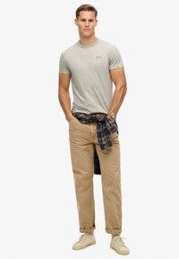 Maglietta grigia in cotone con colletto tondo, pantaloni beige a vestibilità rilassata e sneakers marrone chiaro. Camicia flanella a quadri legata in vita.