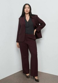Burgundska obleka s prugicami, ki vključuje prilagojen blazer in usklajene hlače. Dopolnjuje jo temno siva majica in črne čevlje z dodatki.