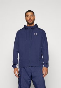Marineblå hættejakke med lynlås, med hvide sideløse og Under Armour-logo, lavet af let, vandafvisende materiale.