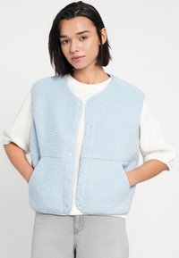 Ανοιχτό γαλάζιο, αφράτο υφασμάτινο γιλέκο με στρογγυλή λαιμόκοψη, κουμπιά με τρύπες και δύο εμπρόσθιες τσέπες, φορεμένο πάνω από λευκό πουλόβερ.