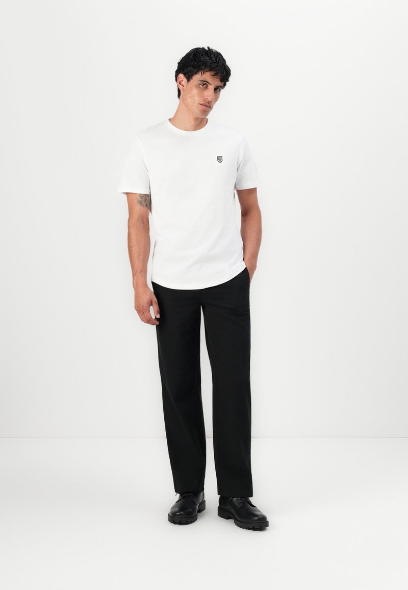 Weißes Baumwoll-T-Shirt mit kurzen Ärmeln, das ein kleines Emblem auf der Brust hat. Getragen mit schwarzen, taillierten Hosen und schwarzen Schuhen.