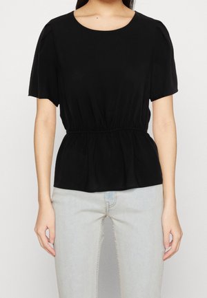 Woman wearing a black short-sleeve blouse with elastic waist and light blue high-waisted jeans, standing against a plain background.