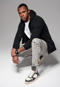 Felpa nera con zip, t-shirt bianca, jeans chiari strappati e sneaker bicolore. Dettagli notevoli includono un orologio da polso e un tessuto testurizzato.