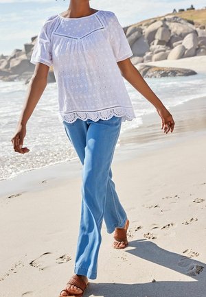 Blusa bianca ricamata con maniche corte, orlo scallop, e jeans di denim blu. Indossata con sandali intrecciati marroni, mentre cammina su una spiaggia di sabbia.