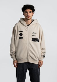 Beige hættetrøje med lynlås og hætte, prydet med forskellige sorte broderede lapper og en afslappet pasform, parret med mørke jeans.
