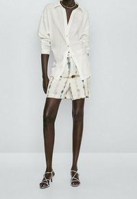 Chemise blanche en lin à boutons, associée à un short tie-dye crème et tons terre, complétée par des sandales argentées à brides.