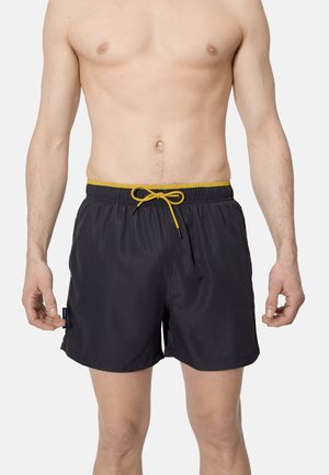 Torso masculino con bañador negro de cintura amarilla y cordón, de pie con los brazos relajados frente a un fondo claro y liso.