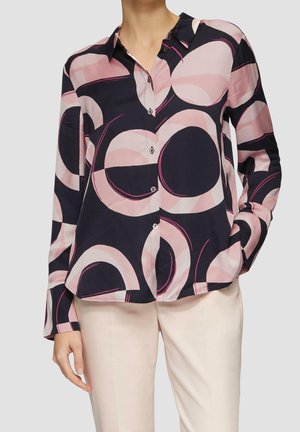 Mujer con blusa negra de botones decorada con grandes patrones circulares en rosa y beige, combinada con pantalones beige claro, sobre un fondo neutro.