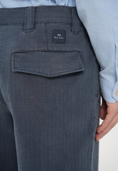 Τα παντελόνια από σκουρόχρωμο τζιν διαθέτουν μια επίπεδη πίσω τσέπη με μια μαύρη ετικέτα χαραγμένη με το "PS Paul Smith". Το ύφασμα έχει λεία υφή και διακριτικό ξεθώριασμα.