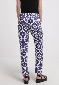 Vita byxor med ett blått geometriskt mönster, med ett elastiskt midjeband och rak benklippning. Modellen har svarta skor med tjocka sulor.