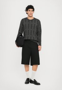 Grauer gemusterter Pullover mit schwarzem Text, schwarze maßgeschneiderte Shorts, weiße Socken und schwarze Slipper. Das Modell hält eine schwarze Stofftasche.