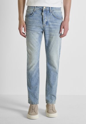 Hombre vistiendo jeans azul claro de pierna recta y zapatillas beige, de pie contra un fondo blanco liso.