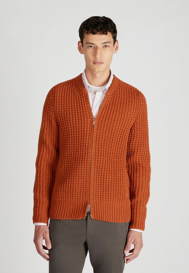 Kardigan z teksturowanej pomarańczowej dzianiny z zapięciem na zamek, prążkowanym kołnierzem i dołem, wyróżniający się dopasowanym fasonem i długimi rękawami noszonymi na białej koszuli.