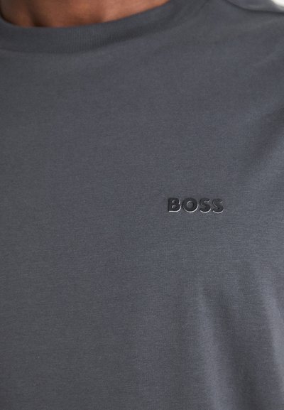 T-shirt gris foncé col rond avec logo noir en relief "BOSS" sur la poitrine gauche, porté par une personne visible du cou vers le bas.