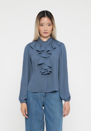 Rynket blå bluse med høy hals, lange ermer og frontspenne, kombinert med lys blå vidde jeans.