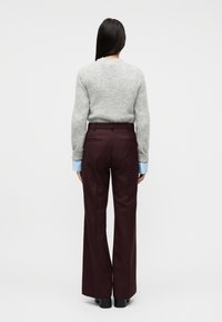 Γυναίκα με μακριά σκούρα μαλλιά που φοράει γκρι πουλόβερ, μανσέτες ανοιχτού γαλάζιου, σκούρα καφέ παντελόνια με φαρδιά πόδια και μαύρα παπούτσια, με την πλάτη προς το μέρος μας.