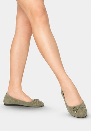 Beine, die olivgrüne Wildleder-Ballerinas mit kleinem Schleifchen und goldenen Spitzen tragen, stehend auf einem neutralen hellgrauen Hintergrund.