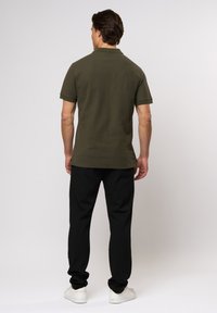 Kaki polo skjorte med korte ermer, ensartet tekstur, sammen med svarte bukser og hvite sneakers. Enkel design, uten synlig maskinvare eller mønstre.
