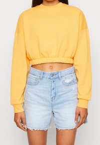 Felpa gialla corta con scollatura rotonda e vita elasticizzata, abbinata a pantaloni corti in denim azzurro con orli sfrangiati.