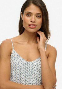 Weißes Camisole mit blauem Blumenmuster, dünnen Trägern, runder Ausschnitt und weichem Stoff. Das Model posiert mit der Hand am Kinn vor einem neutralen Hintergrund.