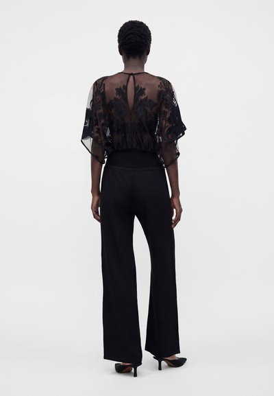 Femme portant un pantalon noir à jambes larges et un chemisier en dentelle florale transparente vue de dos sur fond blanc uni.
